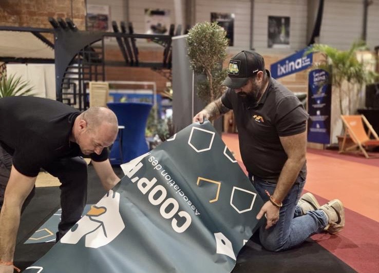 Deux artisans déroulent la bâche Coop’Renov pendant le montage du stand au salon Habitarn 2025